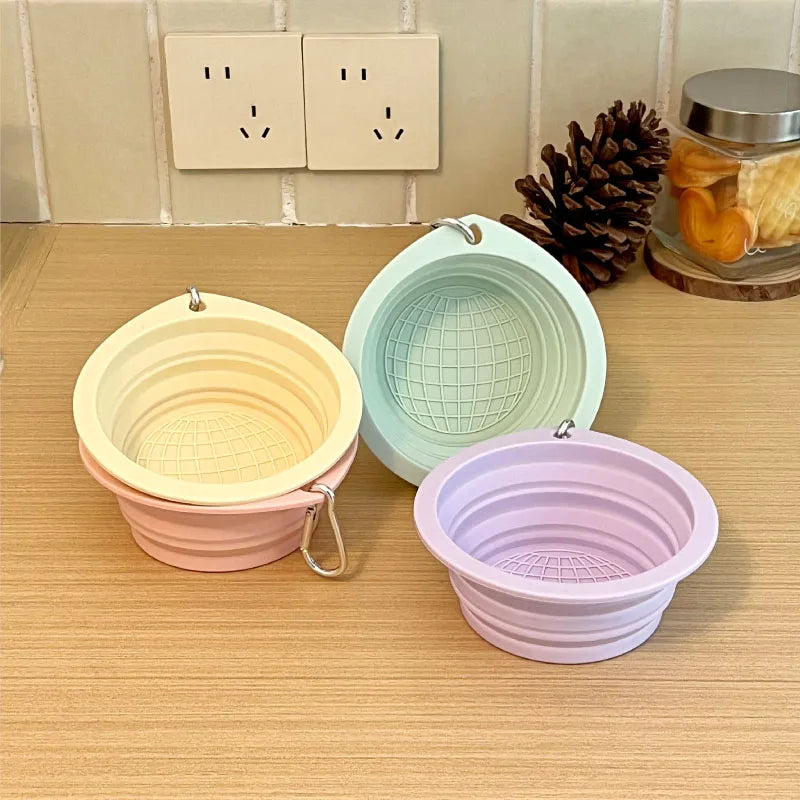 Three collapsible silicone pet travel bowls in pale yellow, mint green, and lavender are displayed on a wooden surface.