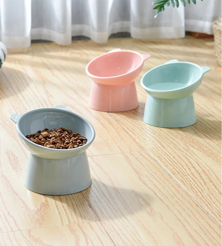 Three ceramic pet feeding bowls in pink and mint green colors resting on a light wooden floor.