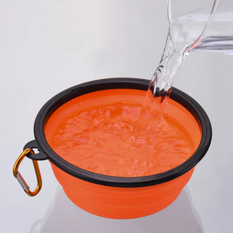 An orange collapsible silicone pet travel bowl being filled with water from a clear glass.