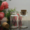 A glass bottle with a cork stopper is shown next to a natural cork, set against a blurred background of pink flowers.