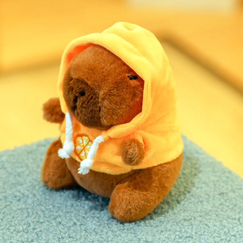Braune Capybara-Plüschpuppe mit leuchtend orangefarbener Kapuzenjacke und weißen Zierleisten und kleinen Flügeln.