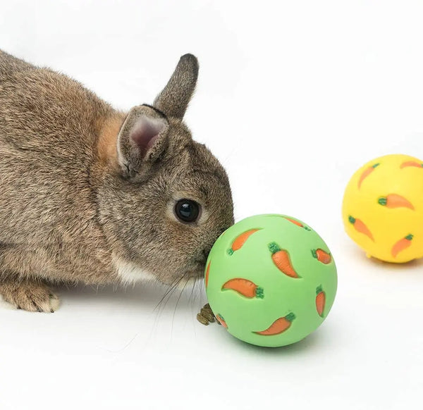 A gray rabbit is sniffing a green ball shaped like a carrot, with a yellow ball nearby.