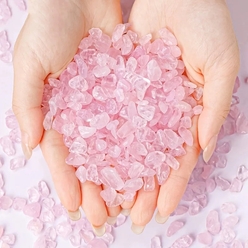 A pair of hands holds a pile of pink rose quartz crystal chips on a white background.