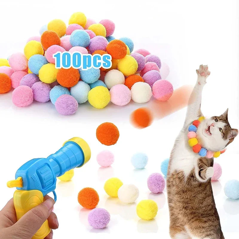 A hand holds a blue plush ball launcher with colorful fuzzy balls scattered around a cat.
