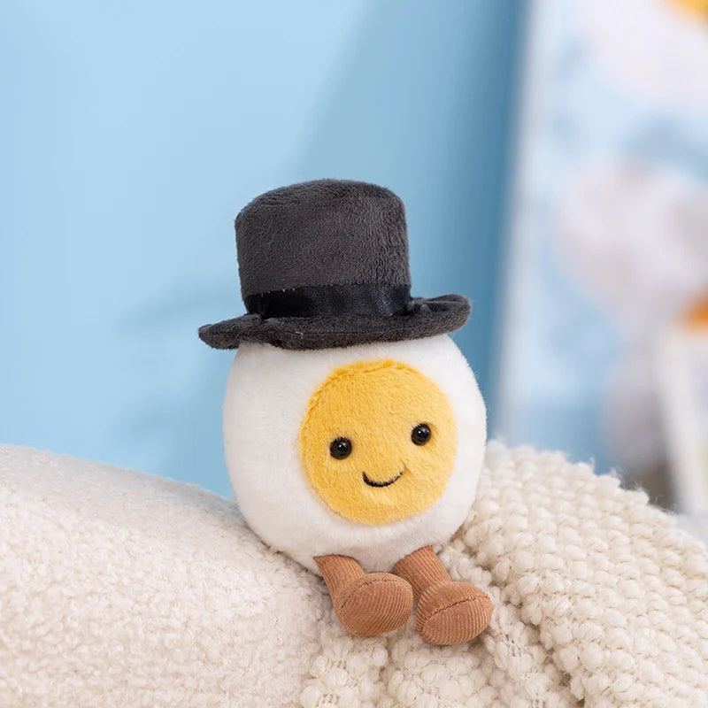 A cute plush toy resembling a sunny yellow egg with a black top and brown feet resting on a cream-colored fabric.