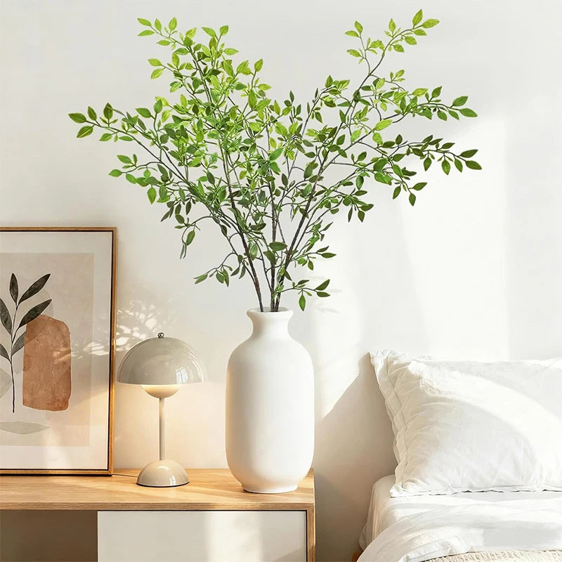 A white ceramic vase filled with green artificial bamboo stems on a wooden shelf next to a lamp and bedding.