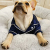 A light-colored dog is wearing a navy blue silk pajama top while lying on a fluffy white surface.