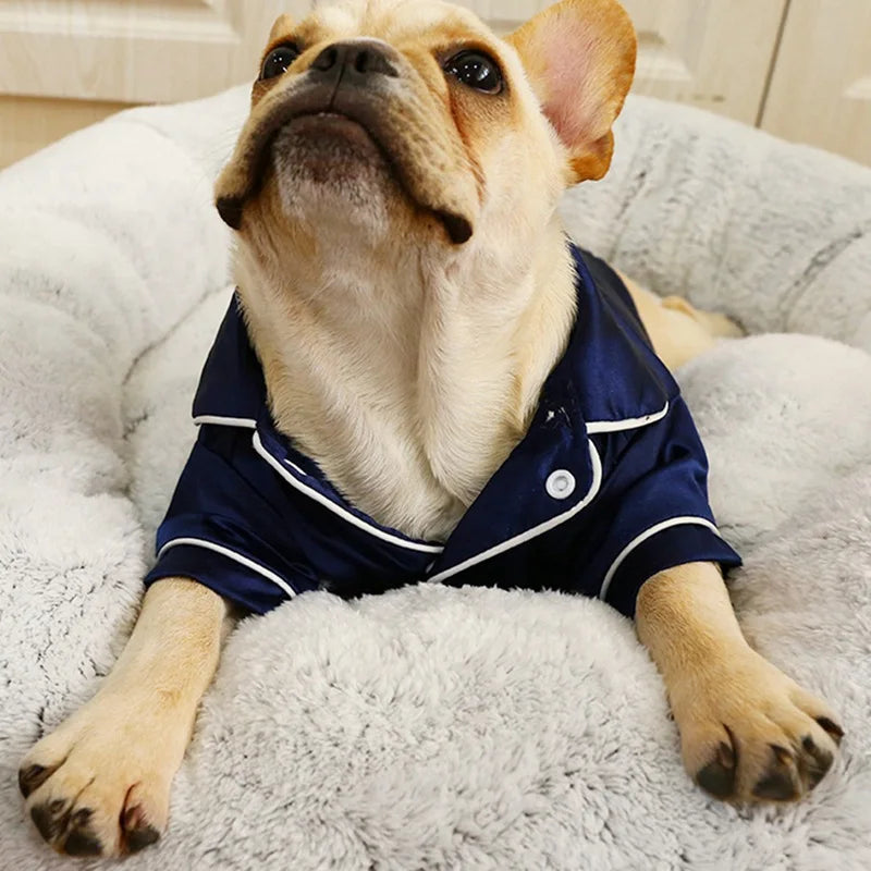 A light-colored dog is wearing a navy blue silk pajama top while lying on a fluffy white surface.