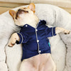 A light-colored dog wearing a navy blue silk pet pajama top, lying on a white fluffy surface.