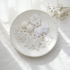 Accessoires pour cheveux en dentelle blanche et pompons, présentés sur une assiette blanche avec une lumière naturelle douce.