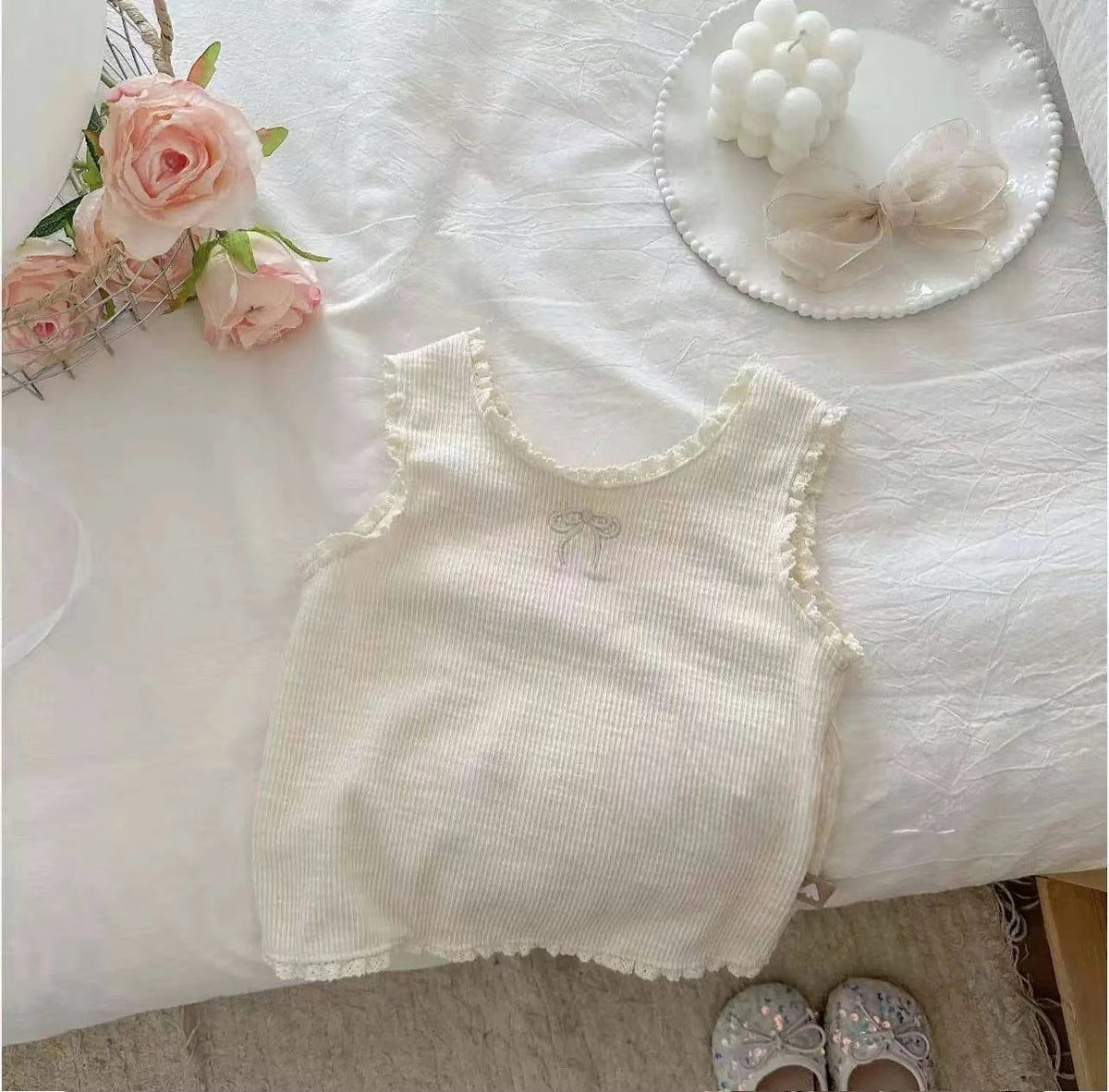 A white mini vest for girls with lace trim, displayed on a white sheet next to pink roses and a bow.