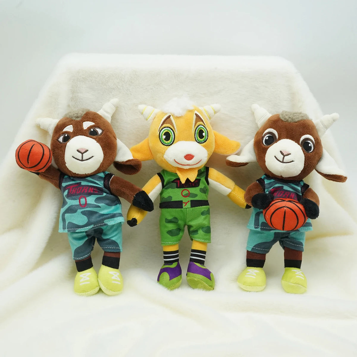 A set of three cute cartoon goat plush toys dressed in sports uniforms sitting on a white fabric surface.