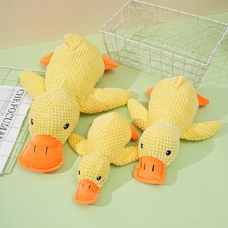 A yellow plush duck dog toy with orange beak and feet is displayed next to a wire basket.