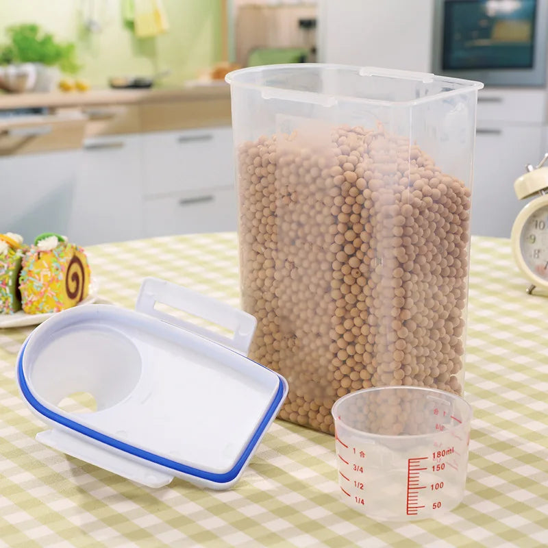 A clear airtight pet food storage container filled with kibble next to a white measuring cup and lid.
