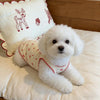 A fluffy white dog wearing a red-trimmed pet tank top, lying on a white surface.