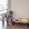A fluffy dog stands next to a wooden feeding stand with two white bowls on a light-colored wall.