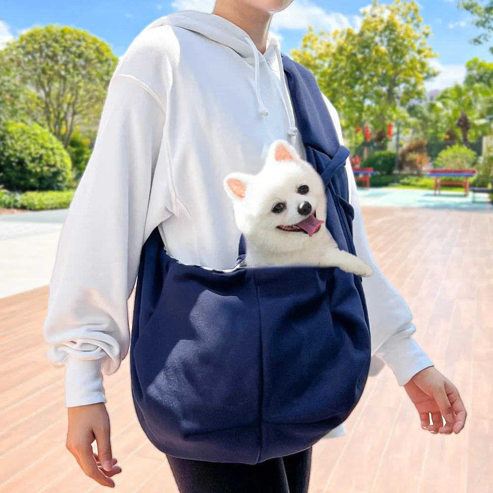 A white hoodie is carrying a small white dog in a navy blue hands-free pet sling carrier.