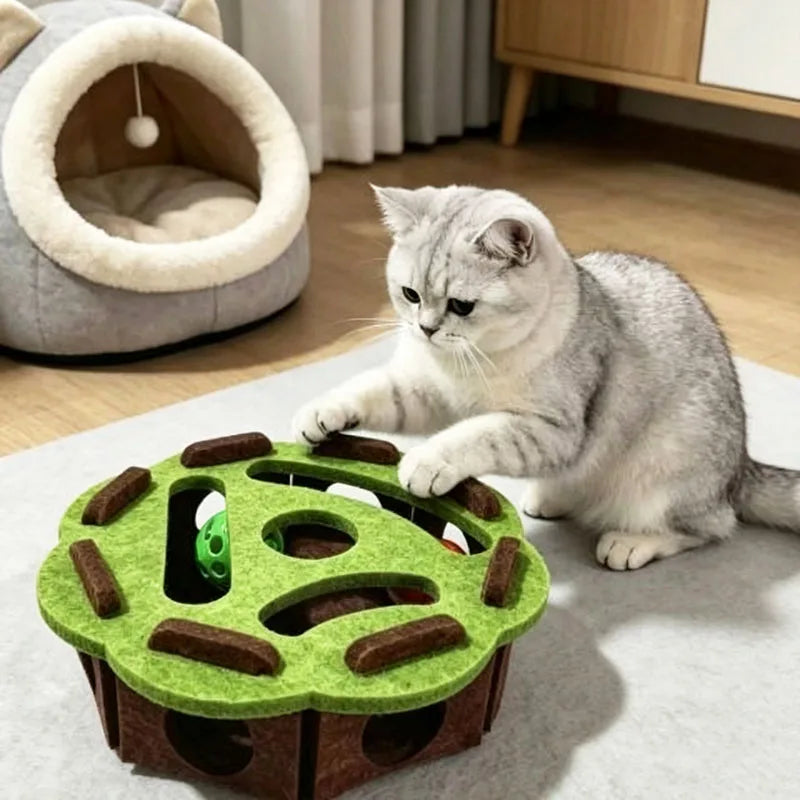 A gray cat is interacting with a green puzzle toy box designed for cats, featuring a maze tunnel and a bell ball.