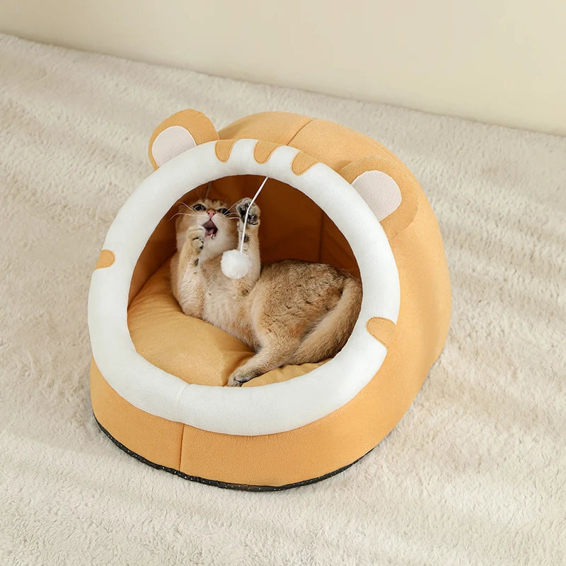 A fluffy, light brown cat is resting inside a round, beige cat-shaped cave bed with a white rim.