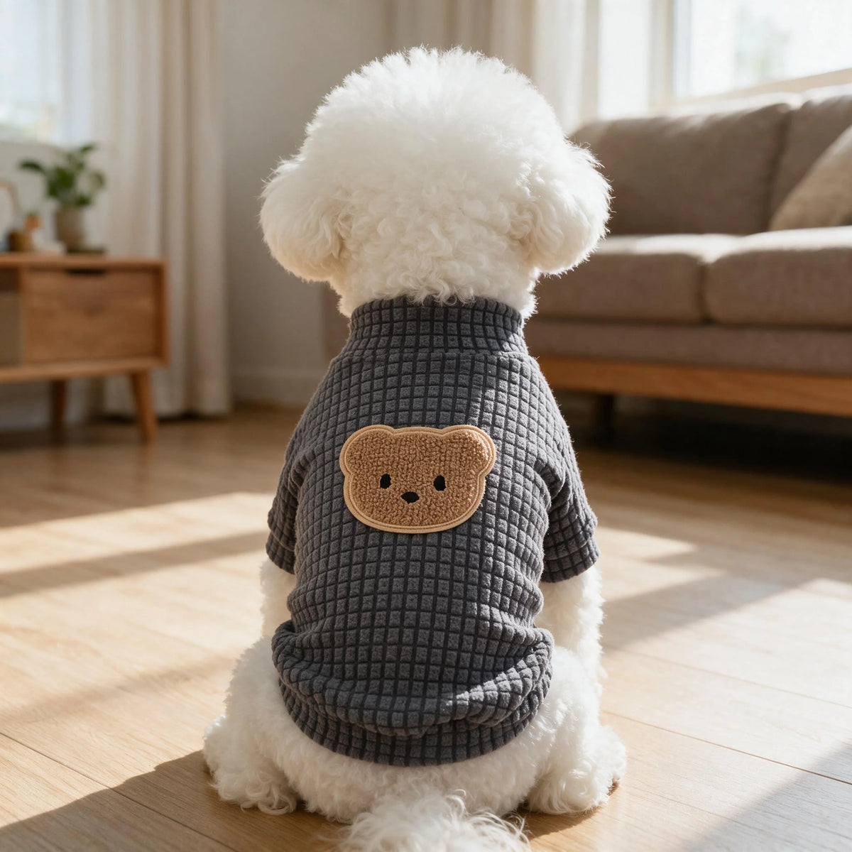 A white dog wearing a gray waffle knit t-shirt with a brown bear embroidery, sitting on a wooden floor.