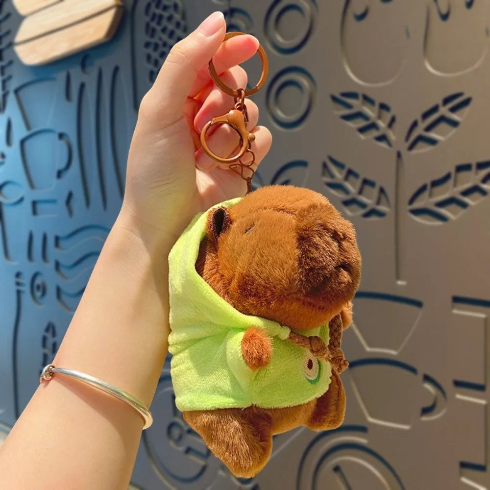 Eine Hand hält eine braune Capybarapuppe mit limegrüner Kapuzenjacke und goldenen Ohrringen vor einem gemusterten Hintergrund