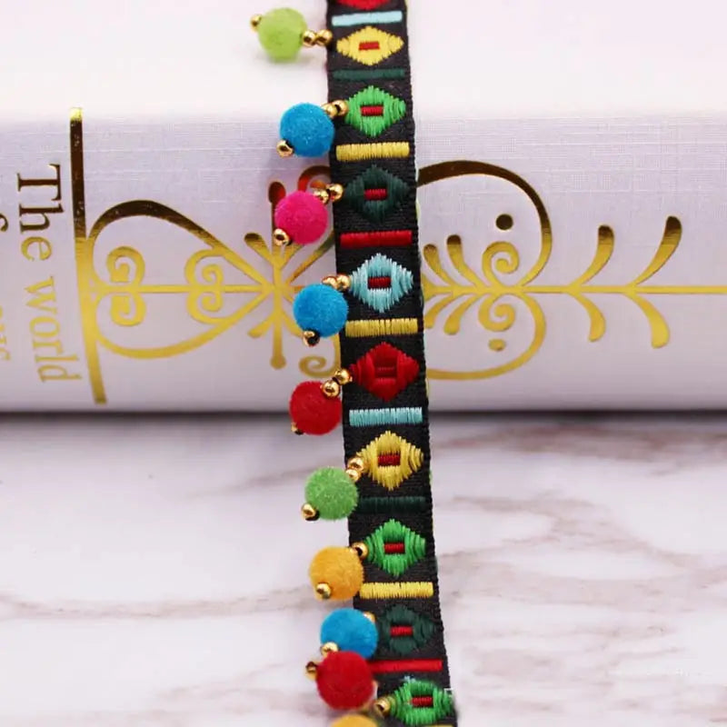 A close-up of a black lace trim featuring colorful pom pom beads in shades of green, blue, pink, and yellow.