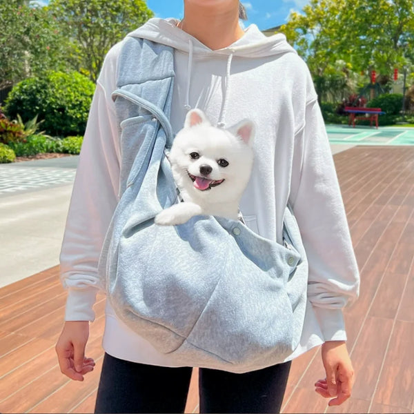 A person wearing a white hoodie is carrying a small white dog in a light blue hands-free pet sling carrier.