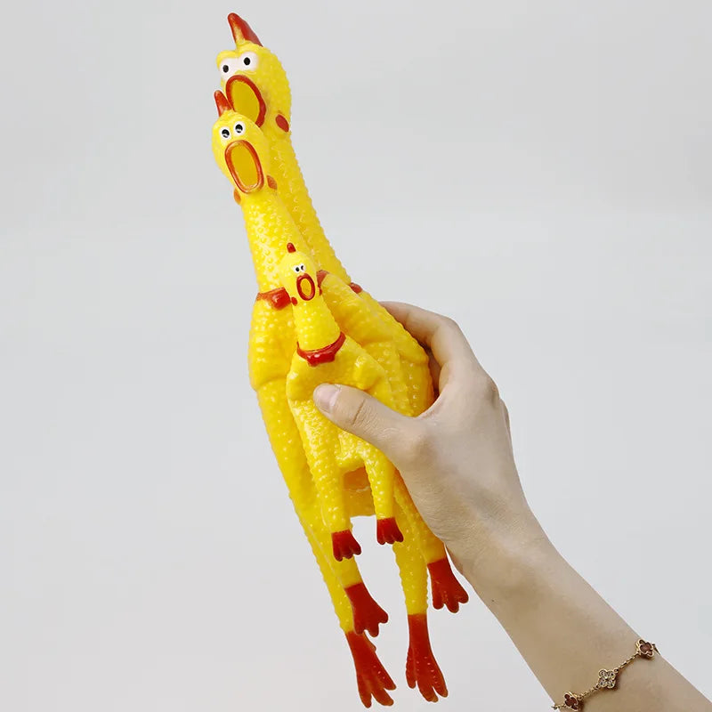 Eine Hand hält einen leuchtend gelben, quietschenden Gummi-Hühner-Hundspielzeug mit orangen Akzenten vor einem hellgrauen Hin