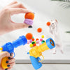 A hand holds a blue toy gun, firing soft orange and purple plush balls toward a white kitten.