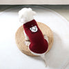 A white dog wearing a red knitted sweater with a black bear design, resting on a woven straw mat.