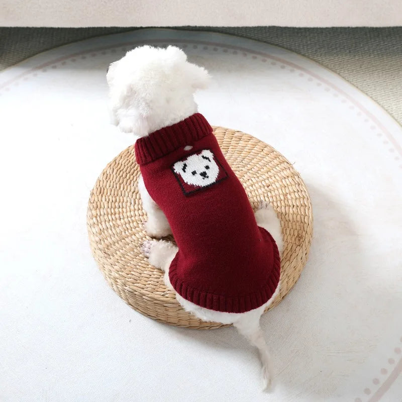 A white dog wearing a red knitted sweater with a black bear design, resting on a woven straw mat.