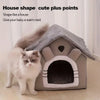 A fluffy white cat sitting next to a gray house-shaped pet bed with an open entrance.