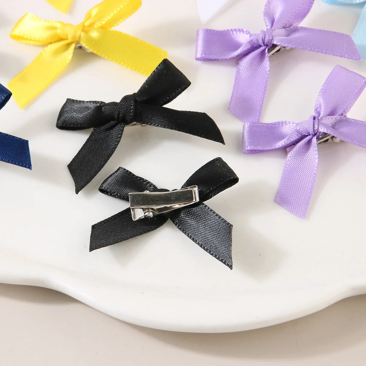 A collection of colorful ribbon bow hair clips in yellow, purple, and blue are displayed on a white surface.