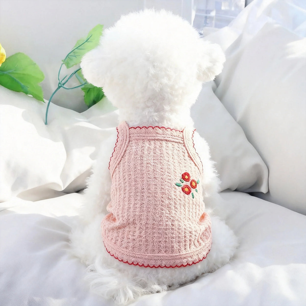 Collar de perro de osito blanco esponjoso con costura rosa y detalle floral rojo, descansando sobre ropa de cama blanca.