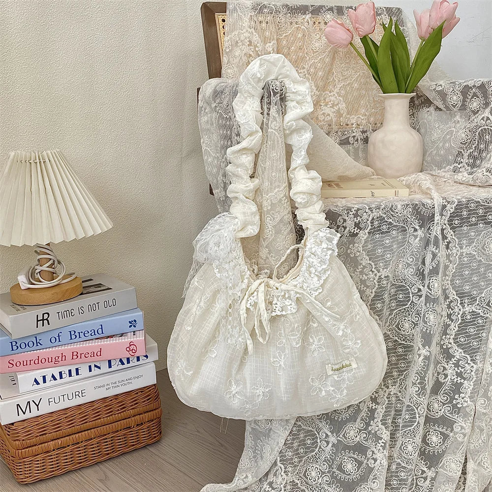 A white floral lace bowknot shoulder bag with a pleated design resting on a textured surface next to books and pink flowers.