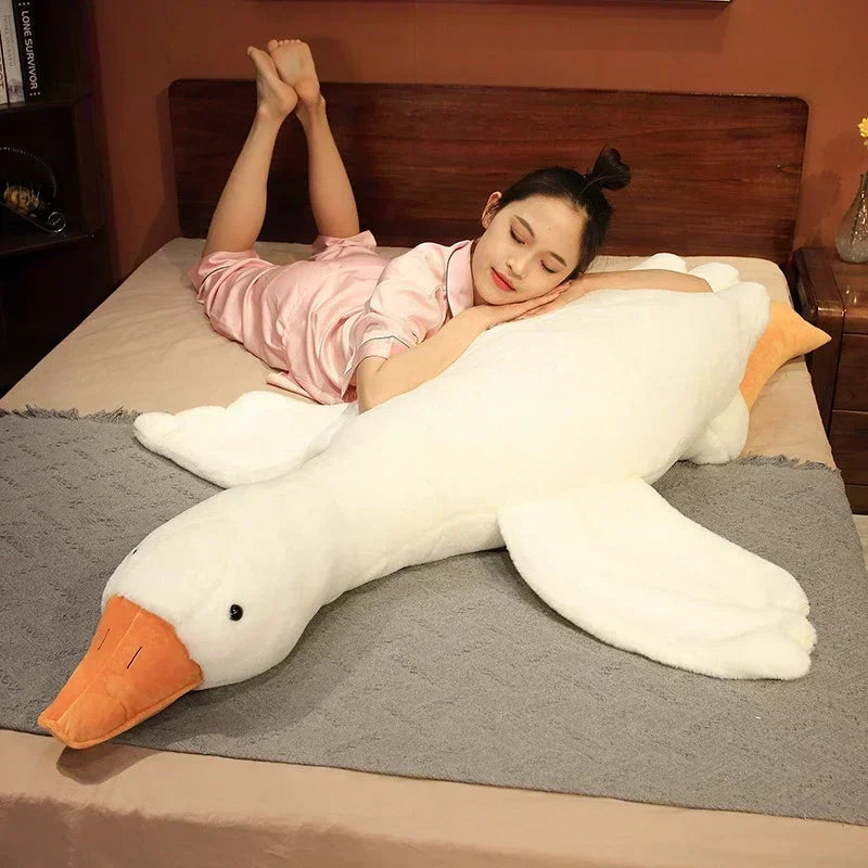 A woman is lying on a bed with a large white plush goose body pillow, looking down.