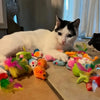 A white and black cat is lying next to a collection of colorful assorted cat toys.