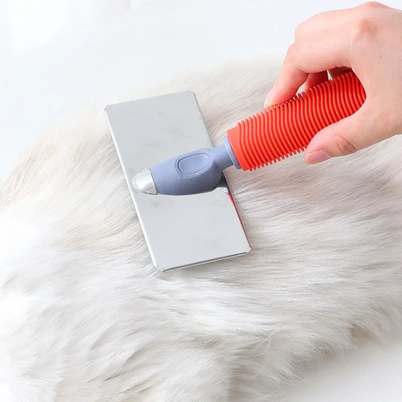 A hand holds an orange silicone cat comb with stainless steel pins over a white fluffy pet.