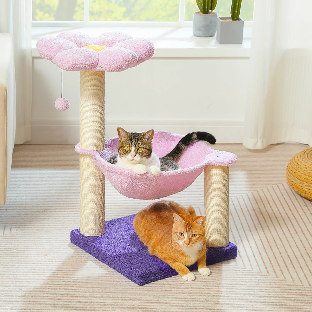 A pink flower cat tree with a hammock and cozy top perch is shown with two cats interacting on the structure.