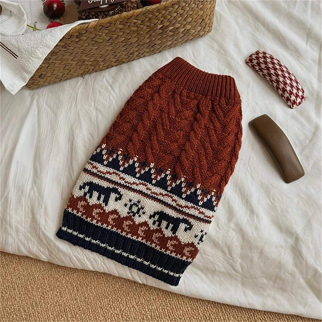 A brown knit sweater with a patterned design is laid on a white sheet next to a woven basket.