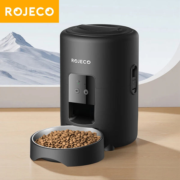A black Rojeco smart pet feeder device sitting on a wooden surface with a bowl of pet food.