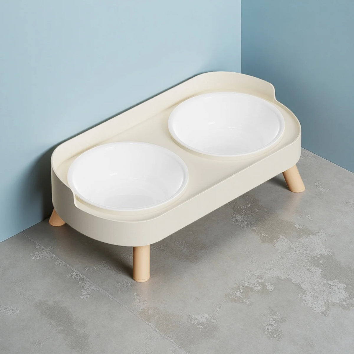 A light beige elevated pet feeder with two white ceramic bowls sits on a gray floor against a blue wall.