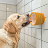 A golden retriever dog is licking a yellow silicone slow feeder bowl mounted on a white tiled wall.