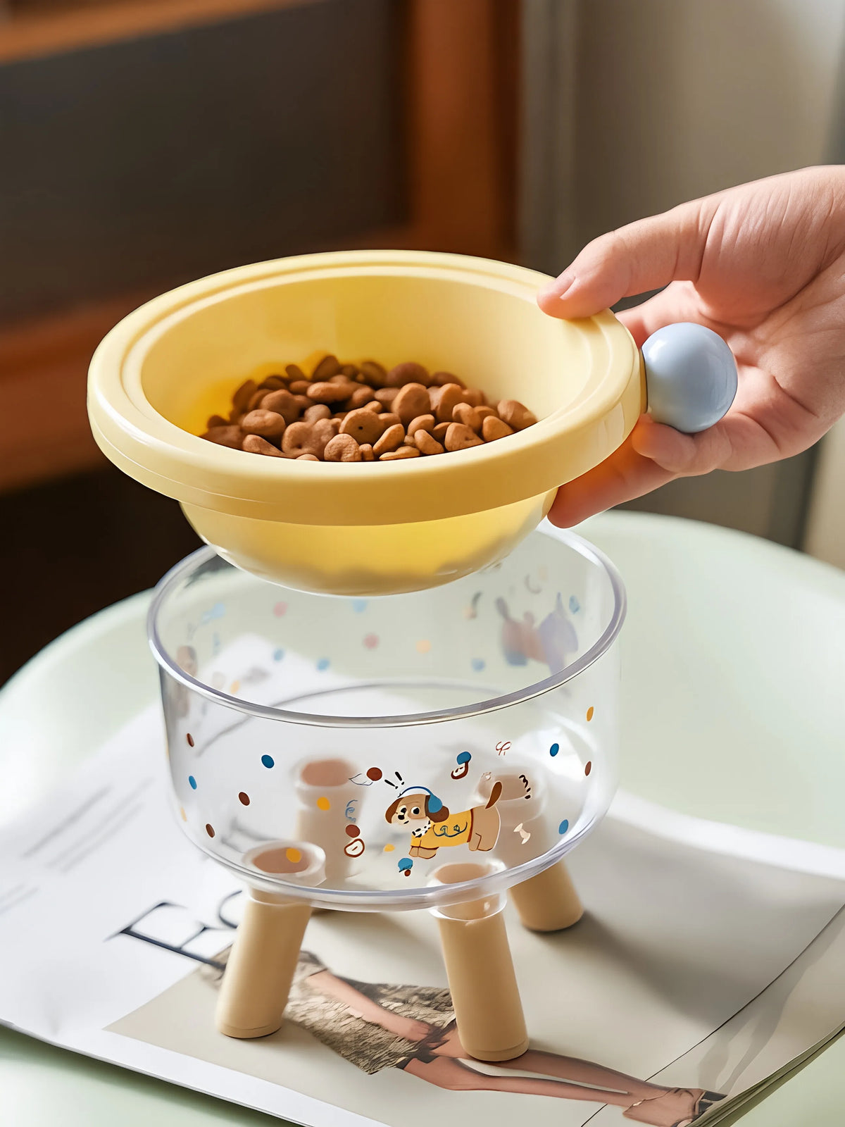 A hand holds a yellow elevated stainless steel double pet bowl with a clear stand, featuring a decorative design.