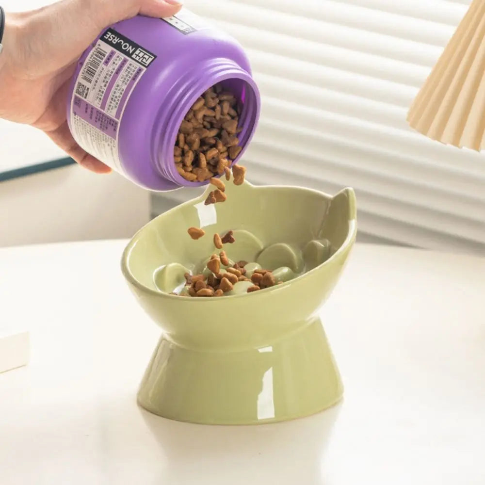 A hand pours brown pet food from a purple container into a light green ceramic bowl.