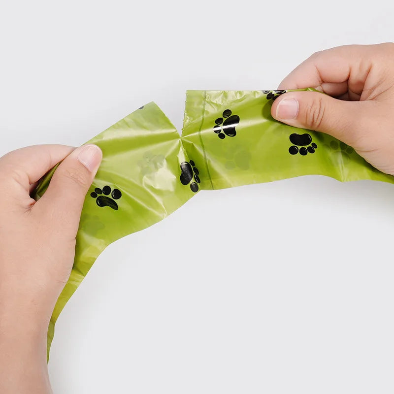 A hand is holding a green biodegradable dog poop bag with black paw prints.