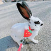 A white cartoon rabbit wearing a red strawberry harness and leash, sitting on an asphalt road.