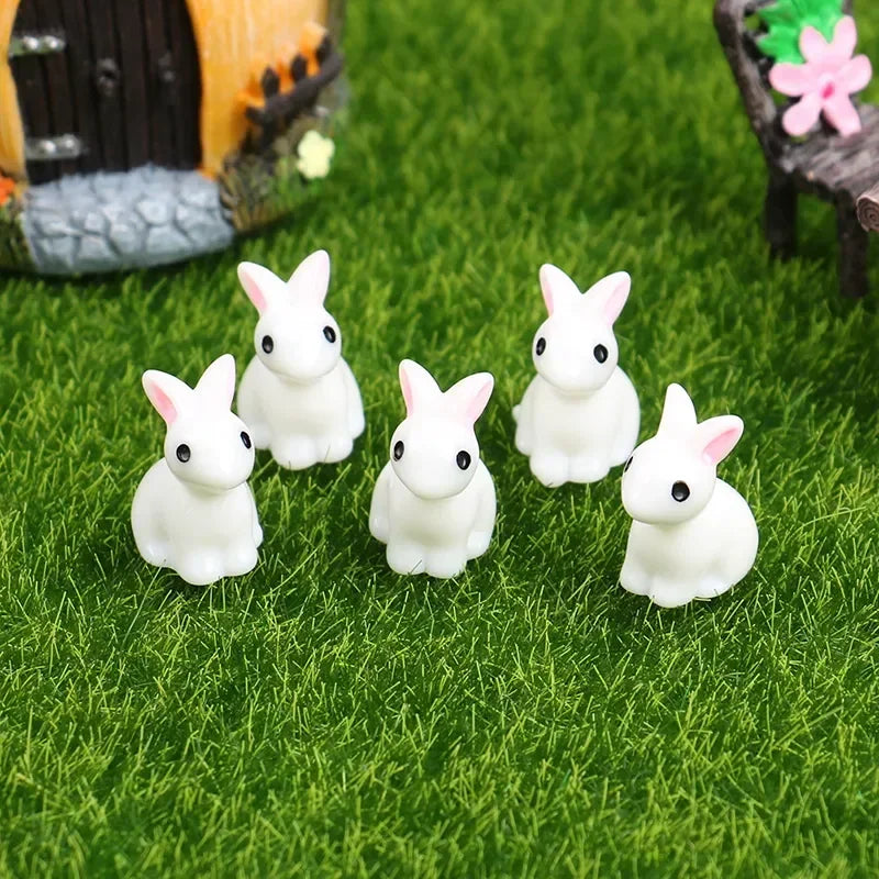 A set of five white resin bunny figurines are displayed on green grass next to a wooden structure.