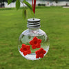 A clear glass bottle filled with water and several red flowers is hanging from a plant branch.