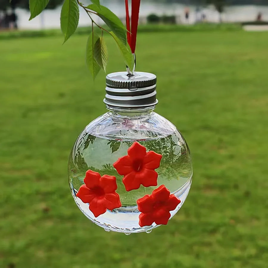 A clear glass bottle filled with water and several red flowers is hanging from a plant branch.